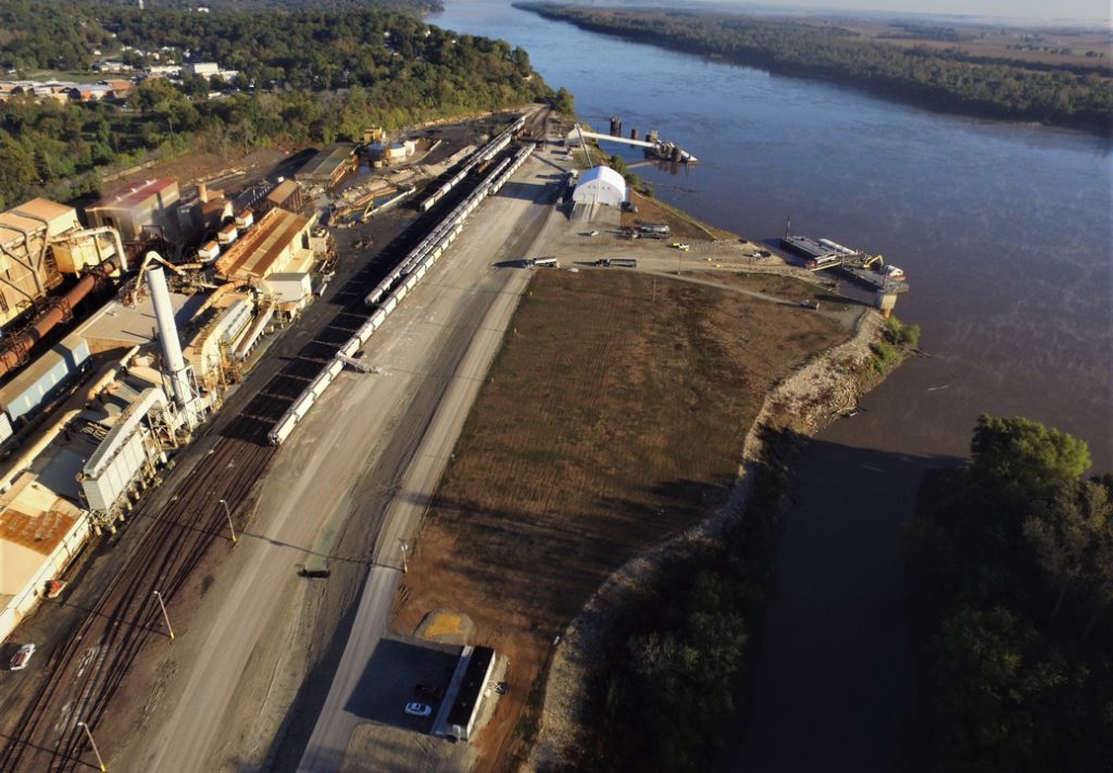 A construction site along a river, featuring several barges moored near the shore. A factory is visible on the riverbank, with smokestacks and industrial structures. The scene captures the interplay between the waterway and the industrial environment, with equipment and materials scattered around, highlighting the active nature of the site. Trees and natural surroundings frame the area, adding contrast to the industrial elements.