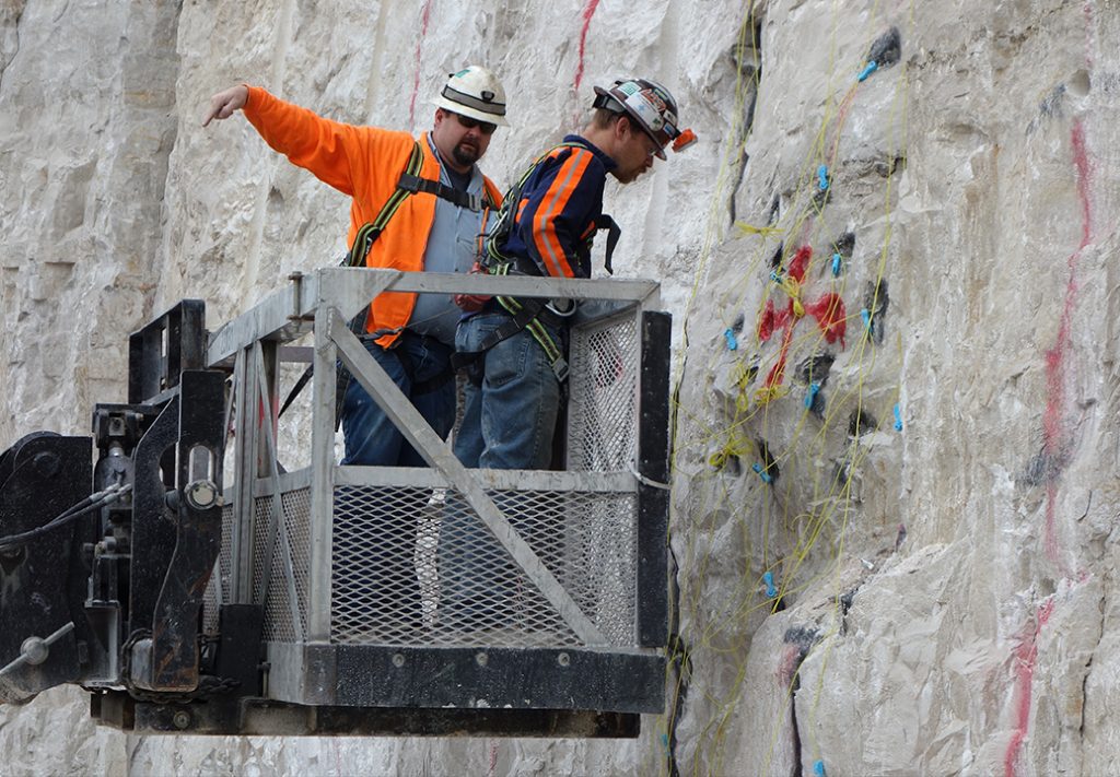 Two workers on a lift inside a mining cave, actively mining the rock walls. They are wearing safety gear and focused on their tasks, using tools to break away material from the sides of the mine. Dim overhead lights illuminate the scene, casting shadows on the rugged rock surfaces, while dust particles float in the air, adding to the industrious atmosphere of the underground environment.