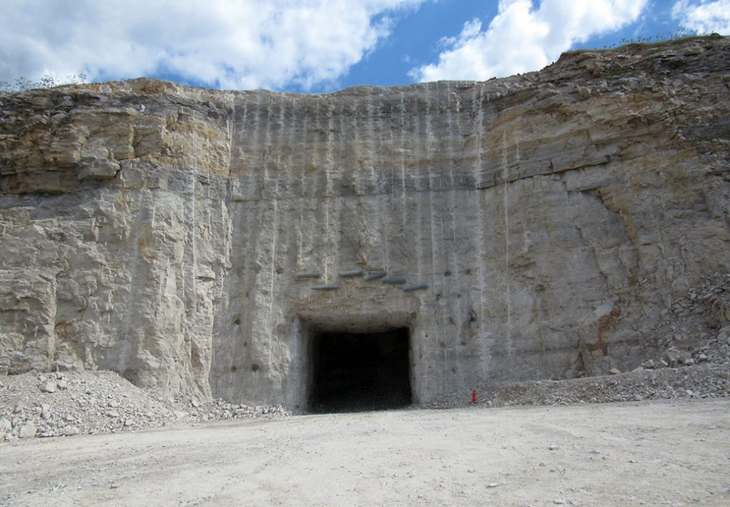 A clear view of the entrance to a cave-like mine, showcasing a large, gaping opening surrounded by rugged rock walls. The ground is littered with loose dirt and scattered rocks, and the entrance is framed by the natural landscape. Sunlight filters in, casting shadows and illuminating the depth of the cave, creating a stark contrast between the bright exterior and the dark interior of the mine.