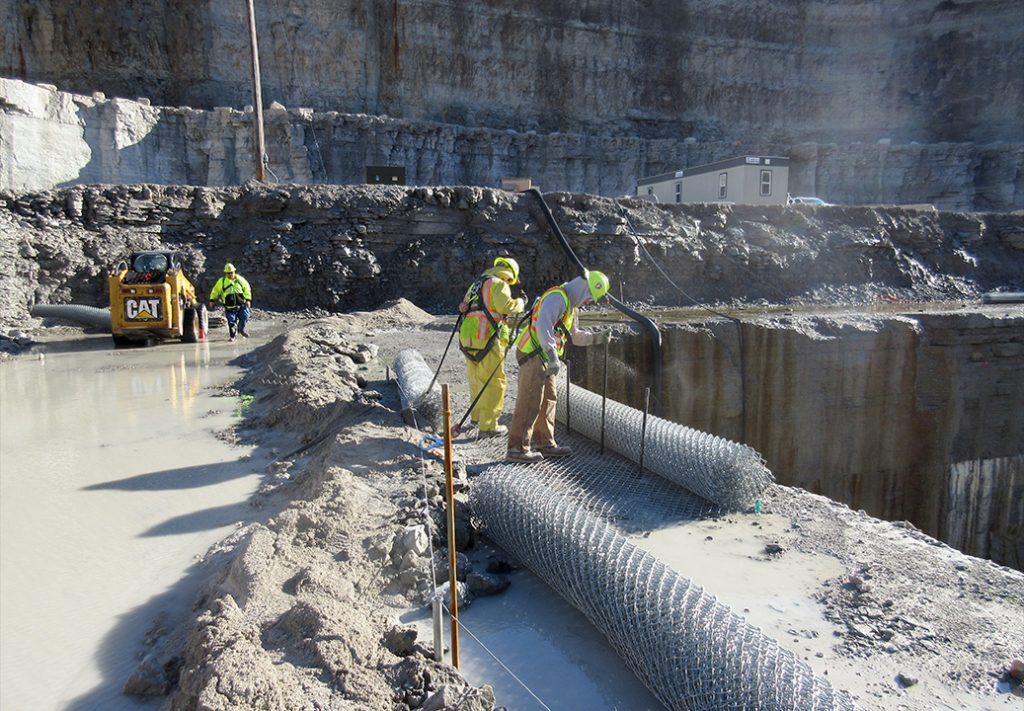 A group of workers standing at the edge of a mining site, near a gaping hole that leads into the mine. A skid steer is parked nearby, ready for use. The workers are wearing safety gear and discussing their next steps, with loose rocks and dirt scattered around them. The rugged landscape and bright sky create a stark contrast to the dark entrance of the mine.