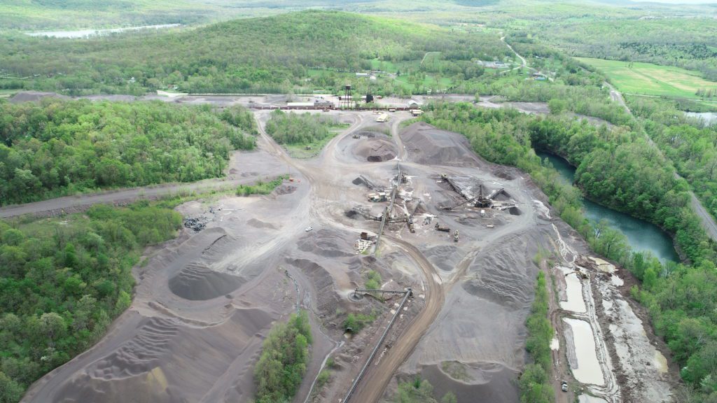 An aerial view of a large mining site, showcasing buildings, machinery, and large piles of excavated rock spread across the area. The site is busy with industrial activity, while the distant background reveals rolling green hills, providing a stark contrast between the rugged, industrial worksite and the natural landscape. The buildings and equipment are strategically placed around the rock piles, emphasizing the scale and scope of the operation.