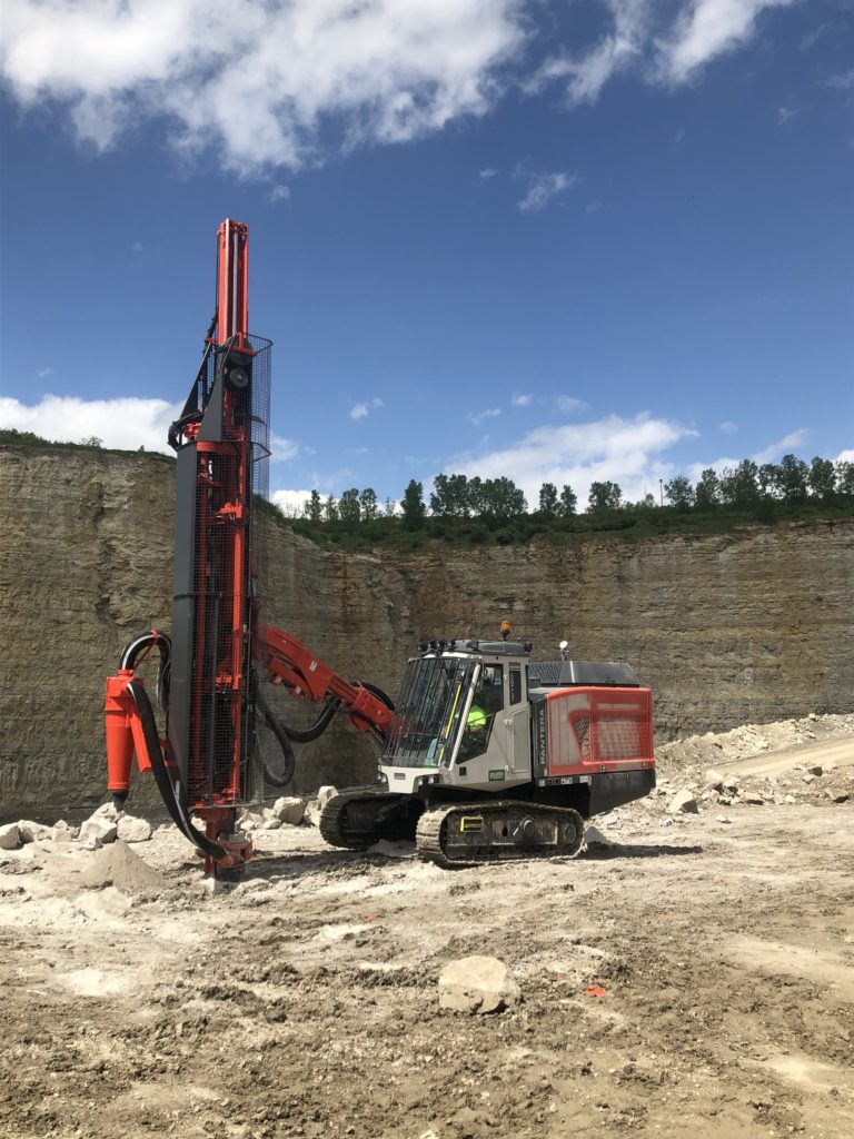 A drilling machine positioned outside of a mining site, ready for operation. The machine is large and robust, equipped with a drill bit at the front, designed for penetrating rock and soil. Surrounding the drilling machine are various tools and equipment typical of a mining operation, with piles of excavated material nearby. The background may include rugged terrain or rock walls, indicating the mining site’s industrial environment. The scene conveys the machine's purpose in facilitating extraction or exploration activities. 4o mini