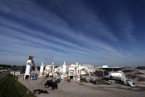 The image depicts a large industrial quarry or mining site with various conveyor belts, crushers, and processing machinery. Sand or gravel piles can be seen around the equipment, suggesting that the site is involved in the extraction or processing of aggregates. There are also several containers, vehicles, and a water truck visible, indicating an active, large-scale operation.